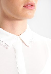 Blusa bianca con colletto classico, caratterizzata da delicati dettagli in pizzo vicino al colletto. Tessuto morbido e trasparente con chiusura a bottoni.