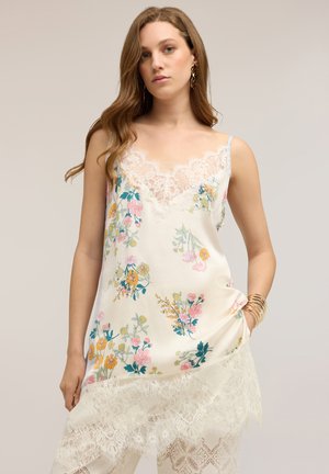 Giovane donna che indossa un vestito a fiori bianco con rifiniture in pizzo, in piedi con una mano in tasca e braccialetti dorati al polso, sfondo neutro.