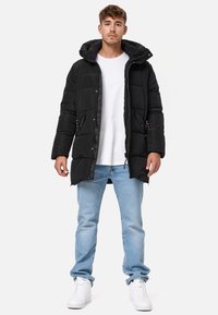 Chaqueta acolchada negra con capucha, con una superficie texturizada, botones a presión y bolsillos laterales. Combinada con jeans azul claro y zapatillas blancas.