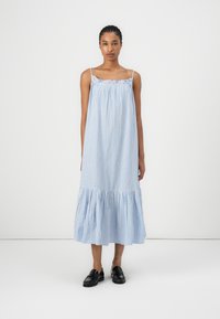 Modrá a biela pruhovaná bezrukávová šaty s zberaným výstrihom, tiered volánkovým lemom a ľahkou tkaninou. Nosené s čiernymi mokasínami.