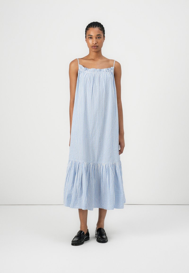 Modrá a biela pruhovaná bezrukávová šaty s zberaným výstrihom, tiered volánkovým lemom a ľahkou tkaninou. Nosené s čiernymi mokasínami.