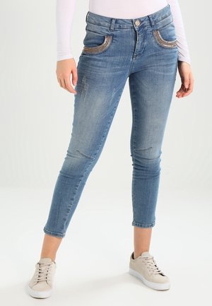 Femme portant un jean skinny court bleu clair avec une bordure décorative aux poches et des baskets beiges, debout devant un fond uni.