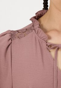 Meka ružičasta bluza s nabranim izrezom, cvjetnim čipkastim detaljima na ramenima, vezanjem i teksturiranom tkaninom.