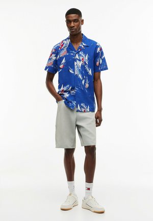 Jeune homme portant une chemise bleue à manches courtes avec des motifs de voiliers, un short gris clair, des chaussettes blanches et des baskets blanches, debout avec une main dans la poche.