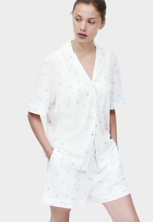 Vrouw draagt een wit overhemd met korte mouwen en knopen en bijpassende korte broek met kleine bloemenprints, handen in de zakken, kijkt naar opzij.
