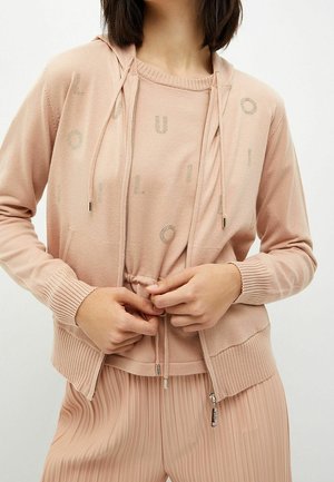 Nainen, jolla on päällään beige vetoketjullinen huppari nyörillä ja siihen sopivat pliseeratut housut, säätää hupun etummaista nyöriä.