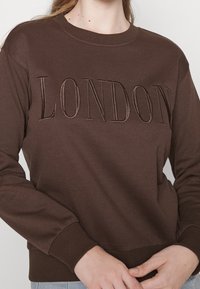 Brun sweatshirt med upphöjda bokstäver "LONDON" på framsidan, ribbade muddar och halsringning samt en mjuk tygstruktur.