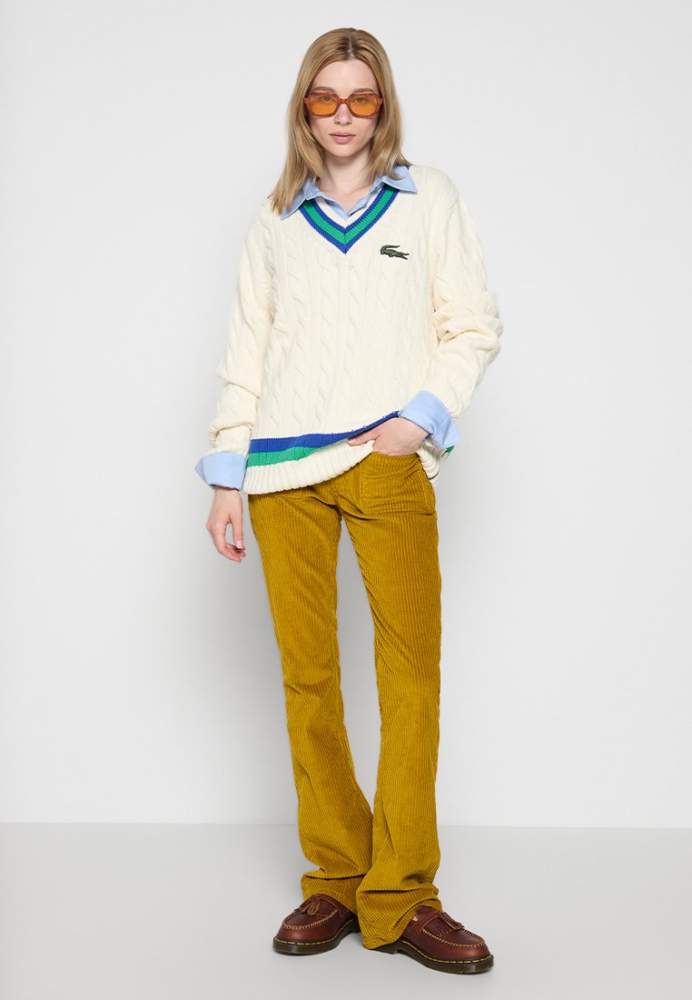 Maglione cable-knit color crema con dettagli a righe multicolori all'orlo, indossato sopra una camicia azzurro chiaro; abbinato a pantaloni in corduroy color senape e scarpe marroni.