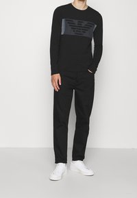 Svart långärmad tröja av jersey med grå panel och logotyp, som är ihop med svarta tapered byxor och vita sneakers mot en neutral bakgrund.