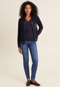 Felpa nera con zip e accenti testurizzati, top scuro con pizzo, jeans blu e mocassini neri. La felpa presenta una tasca frontale.