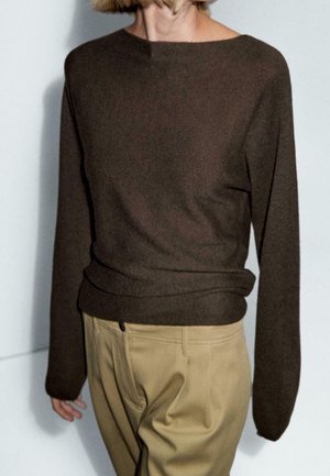 Buste d'une personne portant un pull marron à manches longues et un pantalon beige, debout sur un fond clair uni.