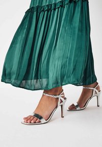 Sandali con tacco alto silver dal design elegante, con cinturini sottili e una texture effetto pelle di serpente, abbinati a un vestito verde plissettato e fluente.