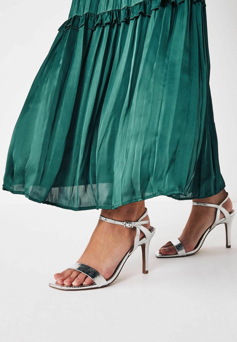 Sandali con tacco alto silver dal design elegante, con cinturini sottili e una texture effetto pelle di serpente, abbinati a un vestito verde plissettato e fluente.
