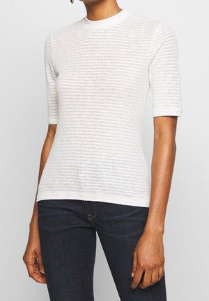 Kvinde iført en hvid, semi-gennemsigtig kortærmet top med vandrette teksturerede striber og mørkeblå tætsiddende jeans.