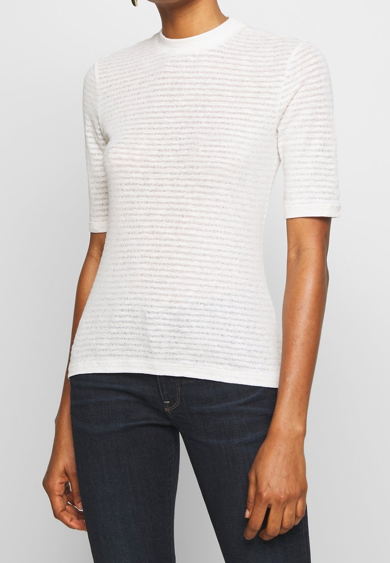 Femme portant un haut blanc semi-transparent à manches courtes avec des rayures horizontales texturées et un jean bleu foncé ajusté.