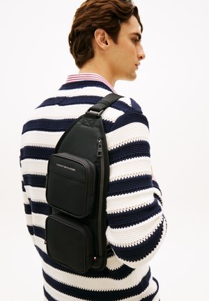 Torba na ramię z czarnego nylonu z dwoma zamykanymi na zamek kieszeniami. Posiada regulowany pasek i detal z logo. Noszona na plecach, na paskowanym swetrze.