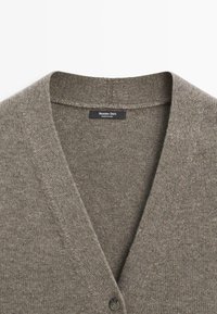 Cardigan en maille gris avec col en V et un bouton visible, portant une étiquette Massimo Dutti à l'intérieur du col.