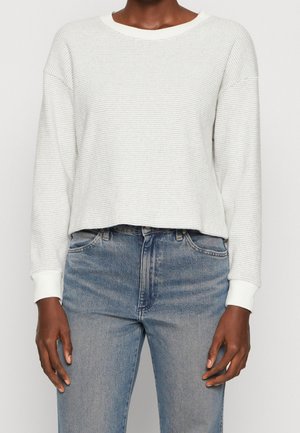 Personne portant un pull blanc texturé à manches longues et un jean bleu taille haute sur un fond uni.
