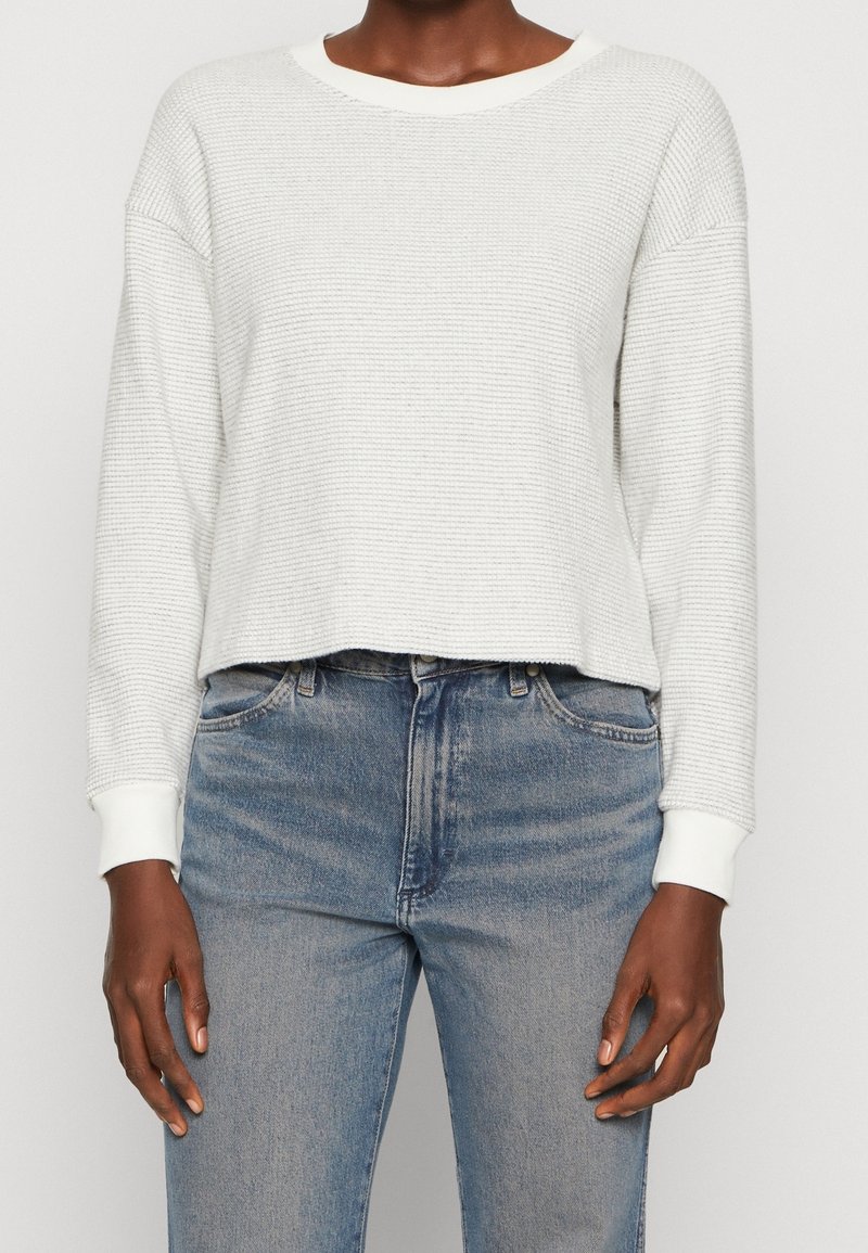 Personne portant un pull blanc texturé à manches longues et un jean bleu taille haute sur un fond uni.