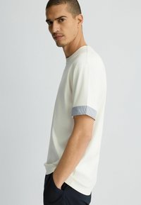 Camiseta blanca de manga corta con cuello redondo y puños a rayas en azul y blanco. Textura suave, corte relajado y detalle en las costuras laterales.