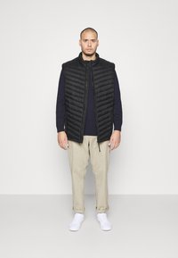 Gilet imbottito nero con design a coste verticali, chiusura con zip, indossato su un maglione blu navy. Pantaloni beige chiaro e sneakers bianche completano l'outfit.