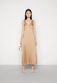 Vestido maxi de cetim bege com decote em V e design sem mangas. Textura suave e silhueta fluida, combinado com sandálias brancas de tiras.