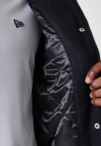 Black coat with a quilted satin interior. Features a white snap button and a small embroidered logo on a light grey shirt.
