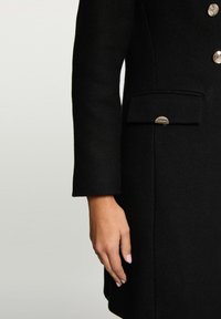 Sort uldfrakke med skræddersyet design, hørmønster, guldfarvede knapper og en sidelomme med en buet metallisk detalje.