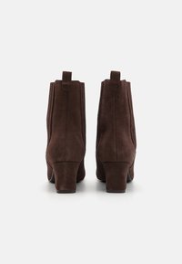 Botas de tobillo de ante marrón con un acabado texturizado, tacón afinado y tiradores en la parte trasera. Presentan un diseño simple y elegante.