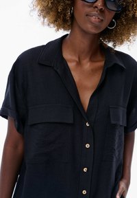 Czarna koszula z krótkim rękawem, wykonana z teksturowanej tkaniny. Posiada kołnierzyk, dwie kieszenie na piersi oraz drewniane guziki na przodzie.