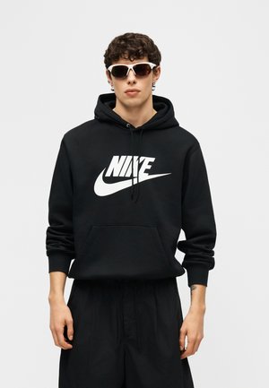 Mlad moški, ki nosi črno Nike kapucar in črne kratke hlače, dopolnjeno z belimi športnimi sončnimi očali, stoji ob preprostem ozadju.