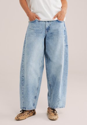 Persoon draagt lichtblauwe wijde jeans, handen in de zakken en bruine gevlekte veterschoenen, staand tegen een effen achtergrond.