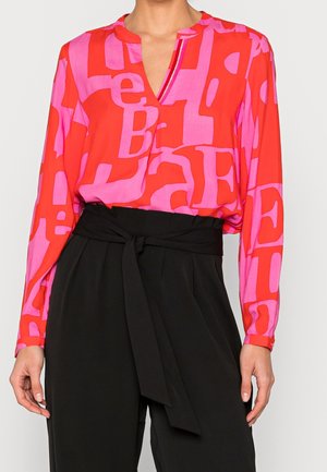 Blouse à manches longues avec col en V, ornée d'un motif abstrait de lettres rouges et roses, rentrée dans un pantalon noir taille haute avec une ceinture large nouée.