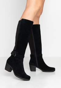 Stivali neri in suede al ginocchio con tacco a blocco e zip laterale, indossati su gambe nude su uno sfondo bianco.