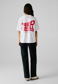 Person med langt mørkt hår iført en hvid, oversized Red Bull Racing t-shirt, sorte bukser og hvide sneakers, stående mod en ensfarvet baggrund.