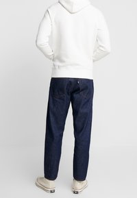 Sudadera blanca con capucha combinada con jeans de denim azul oscuro, que presentan un corte relajado y costuras visibles en los bolsillos traseros. Zapatillas de lona elegantes.