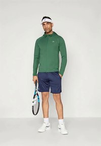 Zaļa sporta jaka ar rāvējslēdzēju, tumši zili šorti un baltas sporta kurpes. Tur rokās tenisa raketi, uzvilkusi baltu vizieri. Vienkāršs dizains un krāsu shēma.