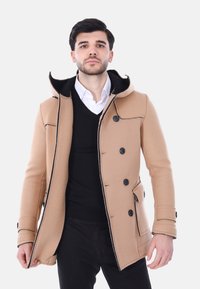Cappotto di lana color sabbia con cappuccio, bottoni neri e bordi neri a contrasto. Abbinato a un maglione scuro e una camicia button-up sottostante.