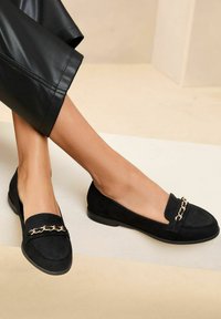 Black suede flats with a chain accent on the upper, rounded toe, and low profile. Smooth texture and minimalist design.