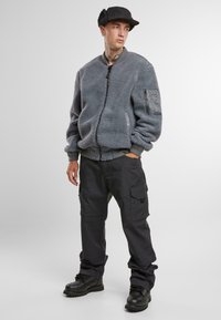 Giacca bomber in pile grigia con chiusura a zip, polsini a coste e una tasca laterale. Indossata con pantaloni cargo neri e stivali neri robusti.