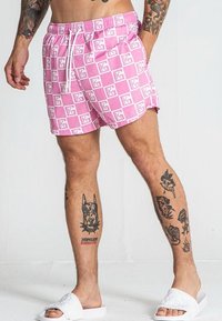 Ružové plavky s bielym geometrickým vzorom, elastickým pásom, sťahovacou šnúrkou a voľným strihom. Nosené s bielymi šľapkami.