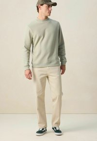 Lichtgroene crewneck sweatshirt, beige broek en donkergroene sneakers. Het item heeft lange mouwen en ribgebreide manchetten bij de polsen.
