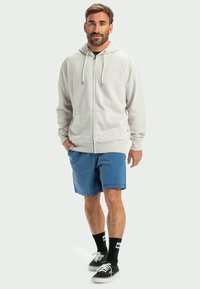 Sweatshirt cinza claro com fecho éclair e capuz ajustável, combinado com calções azuis. Meias e sapatos pretos completam o look casual.