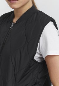 Schwarze gesteppte Weste mit Reißverschluss und geripptem Kragen, mit glatter Textur und ärmellosem Design, über einem weißen Hemd getragen.