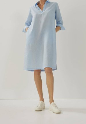 Kobieta w jasnoniebieskiej sukience koszulowej o długości do kolan, z podwiniętymi rękawami i białymi trampkami, stoi na jednolitym tle.