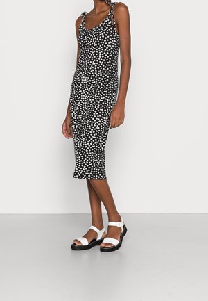 Robe midi noire avec motif floral blanc, bretelles ajustables à nouer et silhouette cintrée. Associée à des sandales blanches avec brides à la cheville.