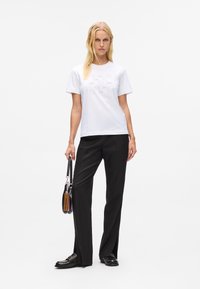 Blonde Frau, die ein weißes Graphic-T-Shirt, schwarze weite Hose, schwarze mit Nieten verzierte Slipper trägt und eine schwarze Handtasche mit Anhängern hält, während sie vor einem weißen Hintergrund steht.