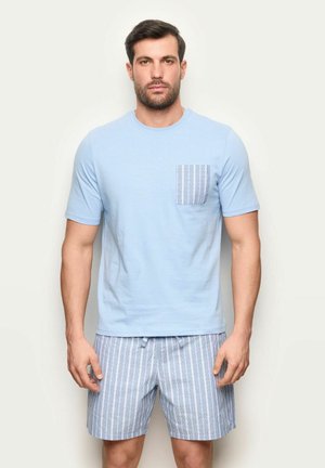 Mężczyzna o krótkich ciemnych włosach, ubrany w jasnoniebieską koszulę z krótkim rękawem z kieszenią w paski oraz pasujące do niej paskowane szorty, stojący na jednolitym tle.