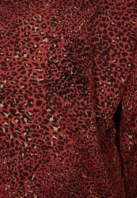 Röd tyg med ett mönster av svart och beige leopardtryck, med en texturerad yta och en liten patchfick på vänster sida.
