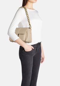 Borsa a tracolla in pelle beige con un design strutturato, caratterizzata da una tracolla in catena dorata e una patta anteriore, appoggiata su una maglietta bianca e jeans scuri.